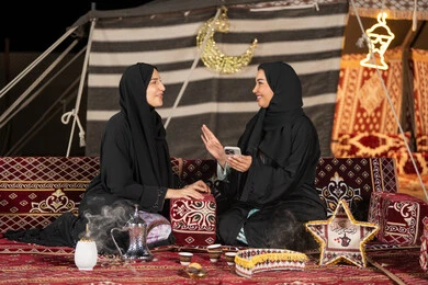 Two smiling Emirati women enjoying talking together, Arab hospitality and reception, use of modern technology, a Saudi Arabian Gulf woman wearing a black abaya sharing online shopping with her friend, gesture of laughter and happiness