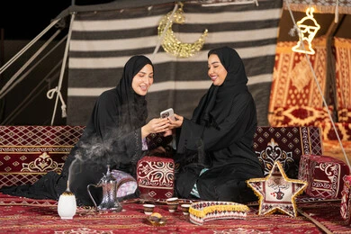 سيدتان اماراتيتان مبتستمتان تستمتعان بالنظر الي الهاتف، حسن الضيافة والاستقبال عند العرب، استخدام التقنيات الحديثه، امرأة عربية خليجية سعودية ترتدي العباءة السوداء تقوم بمشاركه صديقتها في التسوق عبر الانترنت، ايماءه الضحك والسرور