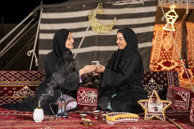 سيدتان اماراتيتان مبتستمتان تستمتعان بالنظر الي الهاتف، حسن الضيافة والاستقبال عند العرب، استخدام التقنيات الحديثه، امرأة عربية خليجية سعودية ترتدي العباءة السوداء تقوم بمشاركه صديقتها في التسوق عبر الانترنت، ايماءه الضحك والسرور