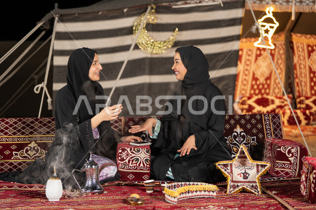 Talking and chatting about happy topics, two Arab Gulf women talking about a topic, using the latest modern technologies, two Saudi women wearing the abaya and hijab sitting in the tent at night, the origins of the popular council