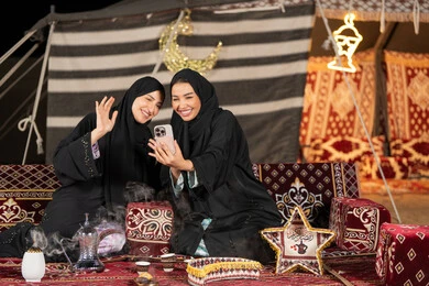 اجراء مكالمه فيديو مع الاهل، سيدتان عربيتان خليجيتان تقومان بالتحدث مع شخص ما, الاطمئنان علي الاهل باستخدام الاتصال عن بعد, استخدام احدث التقنيات الحديثه, امراتين سعوديتان ترتديان العباءه والحجاب تجلسان في الخيمه ليلا، ايماءه الاشاره والترحيب