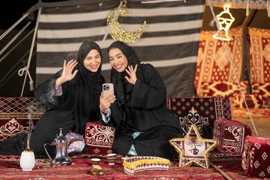 اجراء مكالمه فيديو مع الاهل، سيدتان عربيتان خليجيتان تقومان بالتحدث مع شخص ما, الاطمئنان علي الاهل باستخدام الاتصال عن بعد, استخدام احدث التقنيات الحديثه, امراتين سعوديتان ترتديان العباءه والحجاب تجلسان في الخيمه ليلا، ايماءه الاشاره والترحيب