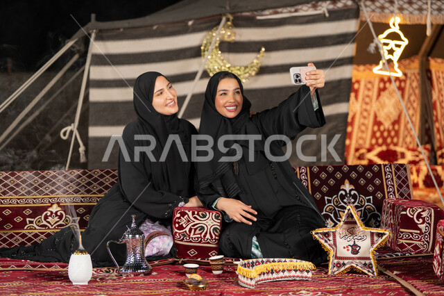 Two Arab Gulf women taking selfies, documenting happy moments with their mobile phones, using the latest modern technologies. Two Saudi women wearing abaya and hijab sitting in a tent at night.