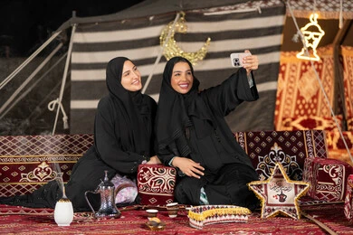 سيدتان عربيتان خليجيتان تقومان بالتقاط صوره سيلفي, توثيق اللحظات السعيده بالهاتف المحمول, استخدام احدث التقنيات الحديثه, امراتين سعوديتان ترتديان العباءه والحجاب تجلسان في الخيمه ليلا