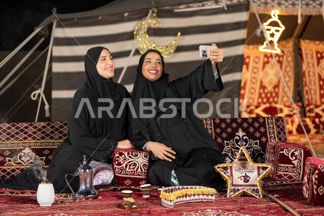 Two Arab Gulf women taking selfies, documenting happy moments with their mobile phones, using the latest modern technologies. Two Saudi women wearing abaya and hijab sitting in a tent at night.