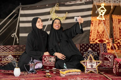 سيدتان عربيتان خليجيتان تقومان بالتقاط صوره سيلفي, توثيق اللحظات السعيده بالهاتف المحمول, استخدام احدث التقنيات الحديثه, امراتين سعوديتان ترتديان العباءه والحجاب تجلسان في الخيمه ليلا