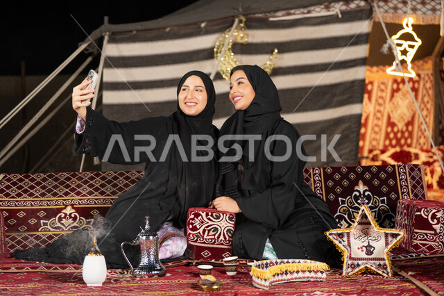 سيدتان عربيتان خليجيتان تقومان بالتقاط صوره سيلفي, توثيق اللحظات السعيده بالهاتف المحمول, استخدام احدث التقنيات الحديثه, امراتين سعوديتان ترتديان العباءه والحجاب تجلسان في الخيمه ليلا