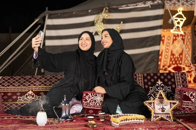 سيدتان عربيتان خليجيتان تقومان بالتقاط صوره سيلفي, توثيق اللحظات السعيده بالهاتف المحمول, استخدام احدث التقنيات الحديثه, امراتين سعوديتان ترتديان العباءه والحجاب تجلسان في الخيمه ليلا
