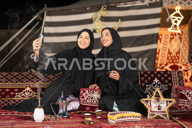 سيدتان عربيتان خليجيتان تقومان بالتقاط صوره سيلفي, توثيق اللحظات السعيده بالهاتف المحمول, استخدام احدث التقنيات الحديثه, امراتين سعوديتان ترتديان العباءه والحجاب تجلسان في الخيمه ليلا