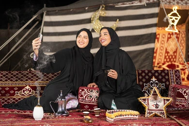 سيدتان عربيتان خليجيتان تقومان بالتقاط صوره سيلفي, توثيق اللحظات السعيده بالهاتف المحمول, استخدام احدث التقنيات الحديثه, امراتين سعوديتان ترتديان العباءه والحجاب تجلسان في الخيمه ليلا