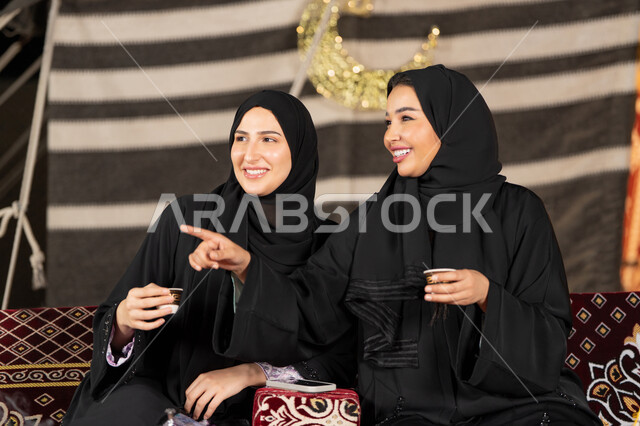 ايماءه الاشاره الي شئ ما، التحدث والتسامر في السوالف السعيده، سيدتان عربيتان خليجيتان تقومان بالنظرالي شئ ما, امراتين سعوديتان ترتديان العباءه والحجاب تجلسان في الخيمه ليلا، اصول المجلس الشعبي، امرأه اماراتيه تشير باصبع السبابه