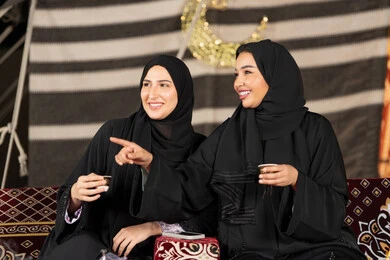 Gesture pointing to something, talking and chatting about happy topics, two Arab Gulf women looking at something, two Saudi women wearing the abaya and hijab sitting in a tent at night, the origins of the popular council, an Emirati woman pointing with her index finger