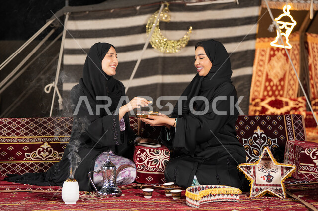 A nutritious breakfast, small fried dough balls, food on the table in Ramadan, good reception and hospitality, an Emirati woman serving appetizers to her friend, a Saudi woman holding a plate full of food