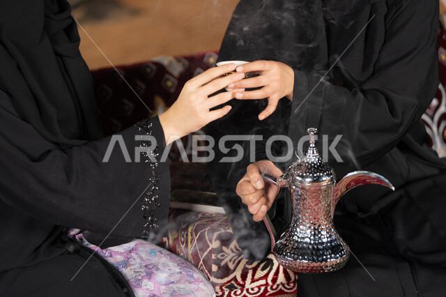 حسن الضيافة والاستقبال عند العرب، العادات والتقاليد الأصيلة في تقديم المشروبات الساخنة،  مفهوم الأنوثة والنعومة، امرأة عربية خليجية سعودية ترتدي العباءة السوداء تقوم بصب القهوة في فنجان لصديقتها، سيدتان اماراتيتان تستمتعان بتناول القهوه