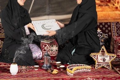 Two Saudi women holding a gift box with the inscription "Mubarak Alaikum Al-Shahr" with a crescent moon and black Ramadan lanterns, congratulations and gift giving in the holy month of Ramadan, an Islamic religious occasion, an Arab Gulf Emirati woman giving a gift to her friend