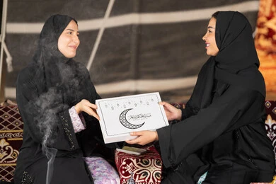 سيدتان سعوديتان تمسكان علبة هدايا عليها مخطوطة مبارك عليكم الشهر مع هلال وفوانيس رمضان باللون الاسود، التهنئه  وتقديم الهديه في شهر رمضان المبارك، مناسبه دينيه إسلاميه، سيده عربيه خليجيه اماراتيه تقدم هديه لصديقتها