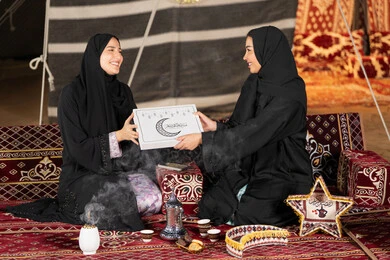 Two Saudi women holding a gift box with the inscription "Mubarak Alaikum Al-Shahr" with a crescent moon and black Ramadan lanterns, congratulations and gift giving in the holy month of Ramadan, an Islamic religious occasion, an Arab Gulf Emirati woman giving a gift to her friend