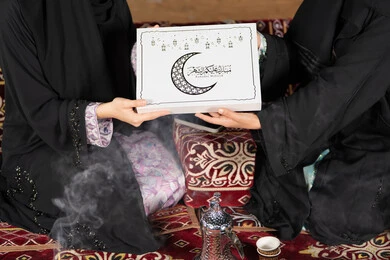 Close-up of two Saudi women holding a gift box with the inscription "Mubarak Alaikum Al-Shahr" with a crescent moon and black Ramadan lanterns. Greetings and gift giving in the holy month of Ramadan, an Islamic religious occasion. An Arab Gulf Emirati woman gives a gift to her friend.