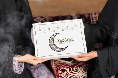 صورة مقربه  ليد امراتين سعوديتان تمسكان علبة هدايا عليها مخطوطة مبارك عليكم الشهر مع هلال وفوانيس رمضان باللون الاسود،  التهنئه  وتقديم الهديه في شهر رمضان المبارك ، مناسبه دينيه إسلاميه، سيده عربيه خليجيه اماراتيه تقدم هديه لصديقتها