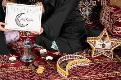 Close-up of two Saudi women holding a gift box with the inscription "Mubarak Alaikum Al-Shahr" with a crescent moon and black Ramadan lanterns. Greetings and gift giving in the holy month of Ramadan, an Islamic religious occasion. An Arab Gulf Emirati woman gives a gift to her friend.