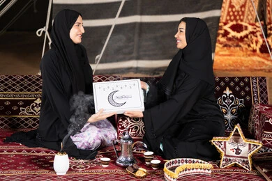 سيدتان سعوديتان تمسكان علبة هدايا عليها مخطوطة مبارك عليكم الشهر مع هلال وفوانيس رمضان باللون الاسود، التهنئه  وتقديم الهديه في شهر رمضان المبارك، مناسبه دينيه إسلاميه، سيده عربيه خليجيه اماراتيه تقدم هديه لصديقتها