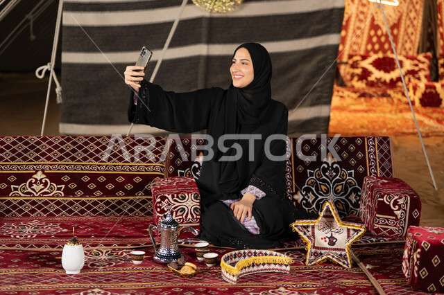 التقاط الصور وتوثيق اللحظات السعيده، سيده اماراتيه ترتدي العباءه السوداء تقوم بالتقاط صوره سيلفي، امرأه عربيه خليجيه سعوديه مبتسمه تقوم باستخدام الهاتف المحمول, المجلس الشعبي التقليدي، التقدم التقني للاجهزه، ديكورات شهر رمضان المبارك
