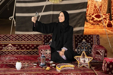 Taking photos and documenting happy moments, an Emirati woman wearing a black abaya taking a selfie, a smiling Saudi Arabian Gulf woman using a mobile phone, traditional folk council, technical advancement of devices, decorations for the holy month of Ramadan