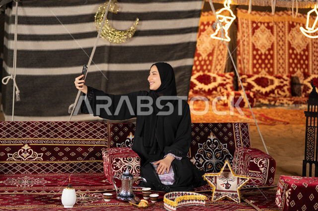 التقاط الصور وتوثيق اللحظات السعيده، سيده اماراتيه ترتدي العباءه السوداء تقوم بالتقاط صوره سيلفي، امرأه عربيه خليجيه سعوديه مبتسمه تقوم باستخدام الهاتف المحمول, المجلس الشعبي التقليدي، التقدم التقني للاجهزه، ديكورات شهر رمضان المبارك