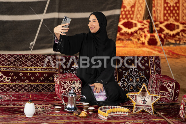 التقاط الصور وتوثيق اللحظات السعيده، سيده اماراتيه ترتدي العباءه السوداء تقوم بالتقاط صوره سيلفي، امرأه عربيه خليجيه سعوديه مبتسمه تقوم باستخدام الهاتف المحمول, المجلس الشعبي التقليدي، التقدم التقني للاجهزه، ديكورات شهر رمضان المبارك