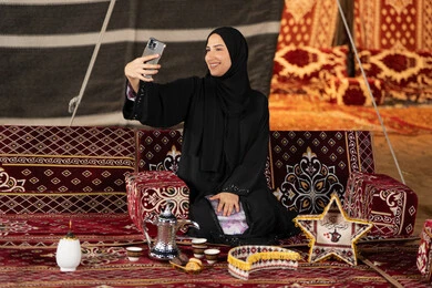 التقاط الصور وتوثيق اللحظات السعيده، سيده اماراتيه ترتدي العباءه السوداء تقوم بالتقاط صوره سيلفي، امرأه عربيه خليجيه سعوديه مبتسمه تقوم باستخدام الهاتف المحمول, المجلس الشعبي التقليدي، التقدم التقني للاجهزه، ديكورات شهر رمضان المبارك