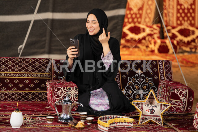 إيماءه الاشاره والترحيب، استخدام الهاتف للدردشه مع الأهل والأصدقاء عبر الانترنت، استخدام التكنولوجيا والتقنيات الحديثة، امرأة عربية سعودية خليجية محجبة تنظرالي الهاتف، سيده اماراتيه تقوم بمكالمه فيديو، استخدام التقنيات والاجهزه الحديثه