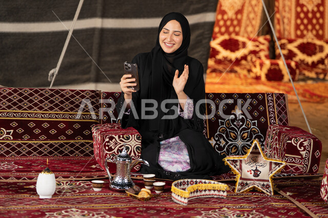 إيماءه الاشاره والترحيب، استخدام الهاتف للدردشه مع الأهل والأصدقاء عبر الانترنت، استخدام التكنولوجيا والتقنيات الحديثة، امرأة عربية سعودية خليجية محجبة تنظرالي الهاتف، سيده اماراتيه تقوم بمكالمه فيديو، استخدام التقنيات والاجهزه الحديثه