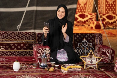 إيماءه الاشاره والترحيب، استخدام الهاتف للدردشه مع الأهل والأصدقاء عبر الانترنت، استخدام التكنولوجيا والتقنيات الحديثة، امرأة عربية سعودية خليجية محجبة تنظرالي الهاتف، سيده اماراتيه تقوم بمكالمه فيديو، استخدام التقنيات والاجهزه الحديثه