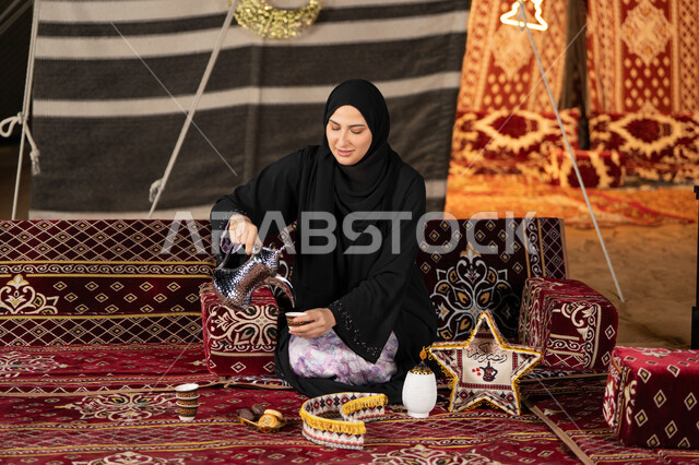 قيم وعادات الضيافه العربيه، حسن الاستقبال وكرم الضيافه،  سيده اماراتيه ترتدي العباءه السوداء تقوم بصب القهوه، امرأه عربيه خليجيه سعوديه مبتسمه تحمل الدله النحاسيه وكوب القهوه, المجلس الشعبي التقليدي