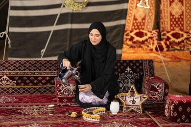 قيم وعادات الضيافه العربيه، حسن الاستقبال وكرم الضيافه،  سيده اماراتيه ترتدي العباءه السوداء تقوم بصب القهوه، امرأه عربيه خليجيه سعوديه مبتسمه تحمل الدله النحاسيه وكوب القهوه, المجلس الشعبي التقليدي