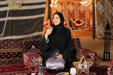 الاستمتاع بتناول القهوه العربيه الساخنه، سيده اماراتيه ترتدي العباءه السوداء تجلس في خيمه وقت الافطار, المجلس الشعبي التقليدي، امرأه عربيه خليجيه سعوديه تمسك كوب القهوه، حسن الاستقبال وكرم الضيافه عند العرب