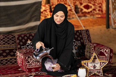 قيم وعادات الضيافه العربيه، حسن الاستقبال وكرم الضيافه،  سيده اماراتيه ترتدي العباءه السوداء تقوم بصب القهوه، امرأه عربيه خليجيه سعوديه مبتسمه تحمل الدله النحاسيه وكوب القهوه, المجلس الشعبي التقليدي