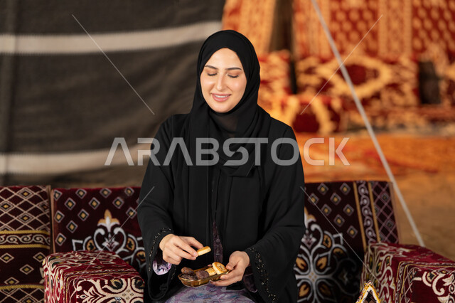 تناول المقبلات اثناء الافطار، امرأه عربيه خليجيه سعوديه مبتسمه تحتفل بقدوم شهر رمضان المبارك, صوره مقربه لسيده اماراتيه ترتدي العباءه والحجاب الاسود تمسك بفانوس معدني ملون, خيمه مليئه بزينه وديكورات رمضانيه, التخييم الليلي الهادئ