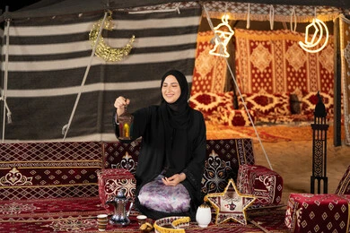 An Arab Gulf Arab Emirati woman sitting in a tent decorated with Ramadan decorations holding a colorful Ramadan lantern, a Saudi woman wearing an abaya and hijab looks with a smile at the metal lantern, decorations for the holy month of Ramadan, the origins of the traditional council in the desert An Arab Gulf Arab Emirati woman sitting in a tent decorated with Ramadan decorations holding a colorful Ramadan lantern, a Saudi woman wearing an abaya and hijab looks with a smile at the metal lantern, decorations for the holy month of Ramadan, the origins of the traditional council in the desert