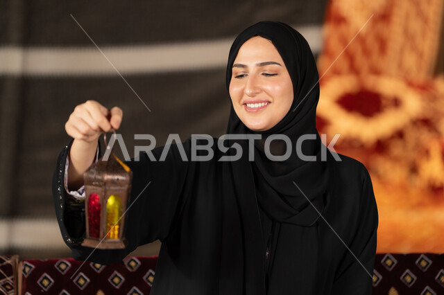 صوره مقربه ليدين امرأه عربيه خليجه عربيه اماراتيه تحمل بيدها فانوس رمضان الملون, سيده سعوديه تقف في خيمه ترتدي العباءه والحجاب تنظر بابتسامه الي الفانوس المعدني, ديكورات شهر رمضان المبارك, اصول المجلس التقليدي في الصحراء