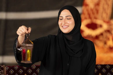 Close-up of the hands of an Arab Gulf Emirati woman holding a colorful Ramadan lantern, a Saudi woman standing in a tent wearing an abaya and hijab looking with a smile at the metal lantern, Ramadan decorations, the origins of the traditional council in the desert