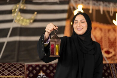 صوره مقربه ليدين امرأه عربيه خليجه عربيه اماراتيه تحمل بيدها فانوس رمضان الملون, سيده سعوديه تقف في خيمه ترتدي العباءه والحجاب تنظر بابتسامه الي الفانوس المعدني, ديكورات شهر رمضان المبارك, اصول المجلس التقليدي في الصحراء
