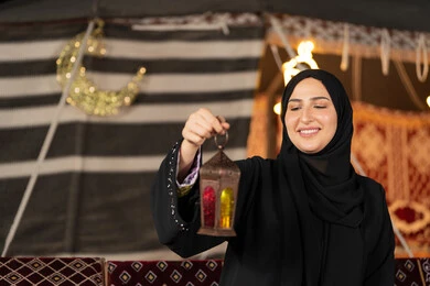 صوره مقربه ليدين امرأه عربيه خليجه عربيه اماراتيه تحمل بيدها فانوس رمضان الملون, سيده سعوديه تقف في خيمه ترتدي العباءه والحجاب تنظر بابتسامه الي الفانوس المعدني, ديكورات شهر رمضان المبارك, اصول المجلس التقليدي في الصحراء