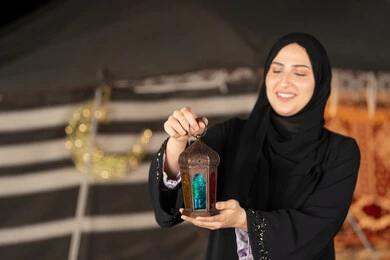 سيده اماراتيه تحمل فانوس معدني, امرأه خليجيه عربيه سعوديه ترتدي العباءه السوداء تمسك فانوس معدني ملون, ايماءه االابتسامه والسرور بقدوم شهر رمضان المبارك, خيمه شعبيه مزينه بديكورات رمضانيه, اصول المجلس الشعبي التقليدي