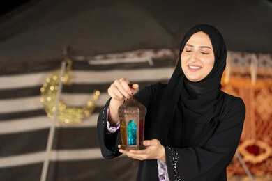 سيده اماراتيه تحمل فانوس معدني, امرأه خليجيه عربيه سعوديه ترتدي العباءه السوداء تمسك فانوس معدني ملون, ايماءه االابتسامه والسرور بقدوم شهر رمضان المبارك, خيمه شعبيه مزينه بديكورات رمضانيه, اصول المجلس الشعبي التقليدي