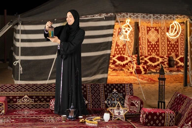 امرأه عربيه خليجه عربيه اماراتيه تحمل بيدها فانوس رمضان الملون, سيده سعوديه تقف في خيمه ترتدي العباءه والحجاب تنظر بابتسامه الي الفانوس المعدني, ديكورات شهر رمضان المبارك, اصول المجلس التقليدي في الصحراء