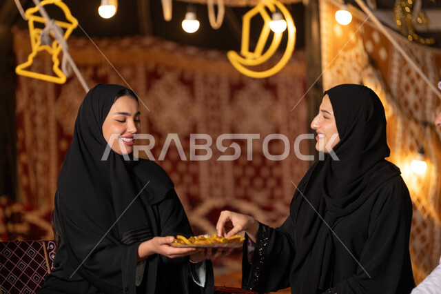 تقديم الحلويات والسكريات في الأعياد والمناسبات في السعودية، صورة مقربة ليد امرأة اماراتيه عربية تمسك بقطعة من الحلوى، حلا عربي شرقي، حسن الاستقبال واكرام الضيف، سيدتان عربيتان خليجيتان سعودتيان ترتديان العباءه الخليجيه يمسكان طبق مليئ من الحلوي