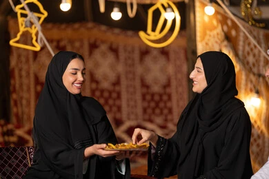 تقديم الحلويات والسكريات في الأعياد والمناسبات في السعودية، صورة مقربة ليد امرأة اماراتيه عربية تمسك بقطعة من الحلوى، حلا عربي شرقي، حسن الاستقبال واكرام الضيف، سيدتان عربيتان خليجيتان سعودتيان ترتديان العباءه الخليجيه يمسكان طبق مليئ من الحلوي