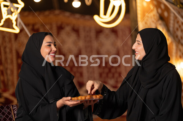 تقديم الحلويات والسكريات في الأعياد والمناسبات في السعودية، صورة مقربة ليد امرأة اماراتيه عربية تمسك بقطعة من الحلوى، حلا عربي شرقي، حسن الاستقبال واكرام الضيف، سيدتان عربيتان خليجيتان سعودتيان ترتديان العباءه الخليجيه يمسكان طبق مليئ من الحلوي