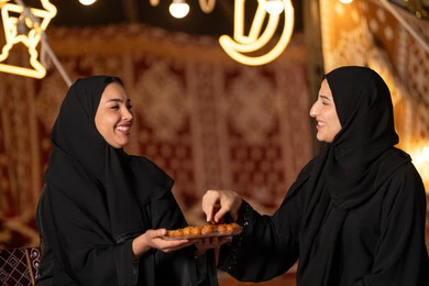 تقديم الحلويات والسكريات في الأعياد والمناسبات في السعودية، صورة مقربة ليد امرأة اماراتيه عربية تمسك بقطعة من الحلوى، حلا عربي شرقي، حسن الاستقبال واكرام الضيف، سيدتان عربيتان خليجيتان سعودتيان ترتديان العباءه الخليجيه يمسكان طبق مليئ من الحلوي